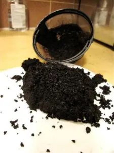 Coffee grounds spilled onto a white plate with a black coffee filter container tipped over in the background.