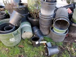 A pile of empty black and green plastic plant pots, some labeled "Monrovia," stacked and scattered on grass near a wall.