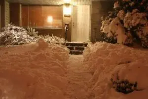 A snow-covered front yard with a shoveled path leading to a lit front door at night. Snow is piled on both sides of the walkway and on bushes.