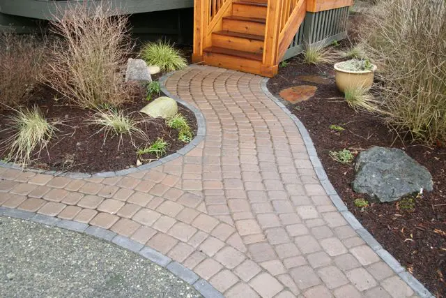 Curved stone walkway with gray border leads to wooden stairs, surrounded by mulch, rocks, small plants, and a potted plant on the right.