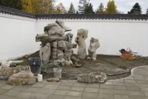 A courtyard with large rocks being arranged, surrounded by a white wall. Construction materials, buckets, hoses, and a wheelbarrow are visible on the unfinished ground.