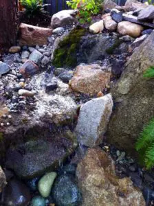 Small garden waterfall flows over rocks and moss, surrounded by stones and green plants.