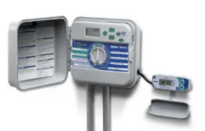 An open irrigation control panel with buttons, a display screen, and a dial, next to a digital timer device attached by a cable.
