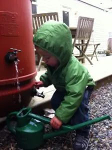 A young child in a green jacket fills a green watering can with water from an outdoor rain barrel beside a wooden deck and patio furniture.