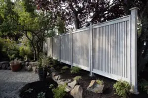 A tall, white vertical slat fence runs alongside a garden with rocks, mulch, small plants, and several trees.