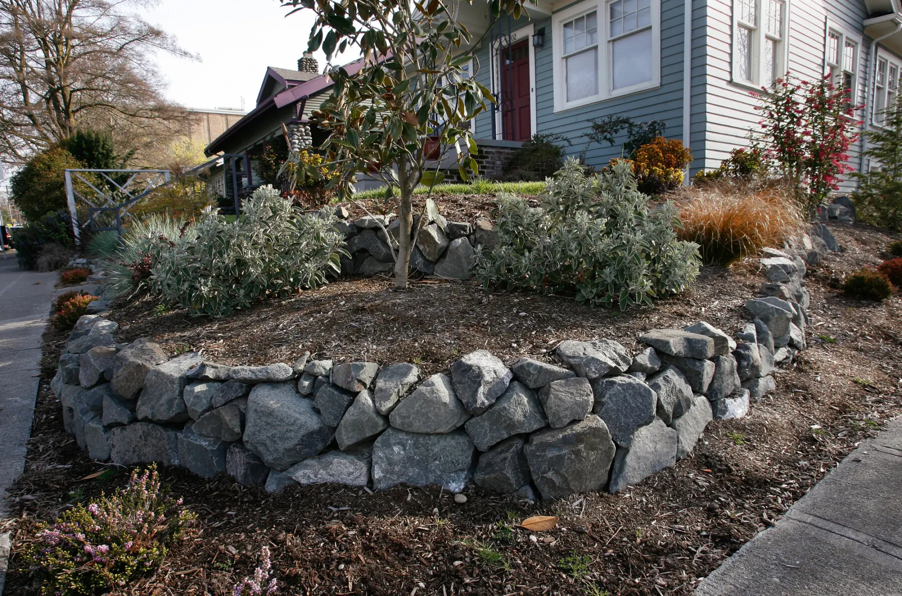 A raised garden bed with stone retaining wall and various shrubs is in front of a gray house with white trim and a red front door.
