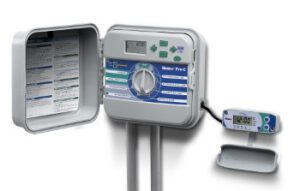 An open irrigation control panel with buttons, a display screen, and a dial, next to a digital timer device attached by a cable.