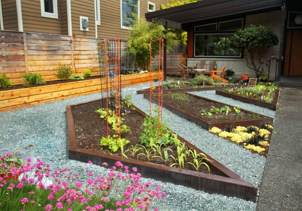 A modern front yard garden with triangular raised beds containing various plants and vegetables, surrounded by gravel paths and a wooden fence.