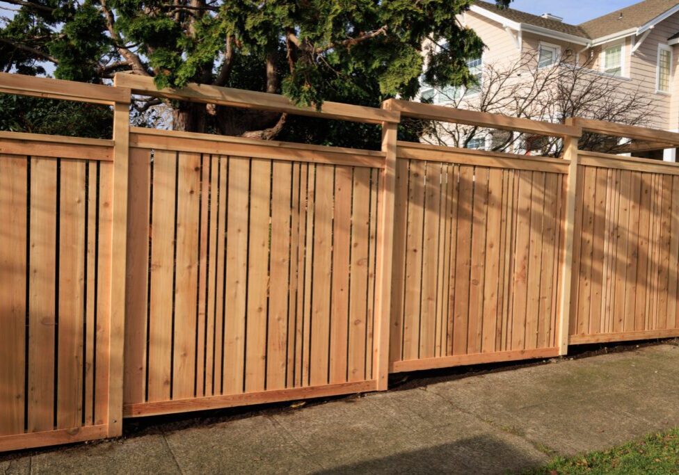 Wooden privacy fence with vertical slats and a horizontal top beam, situated beside a sidewalk in a residential neighborhood.