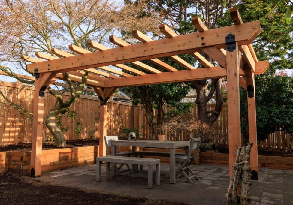 A wooden pergola stands over an outdoor dining table and benches on a stone patio, surrounded by trees and a wooden fence.