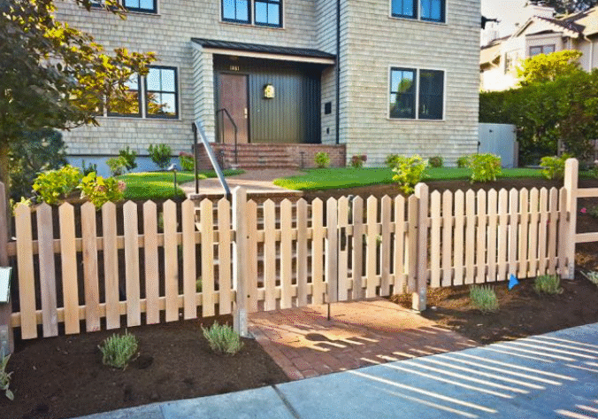 A light wooden picket fence with a gate encloses a front yard, leading to a house with beige siding and a landscaped garden.