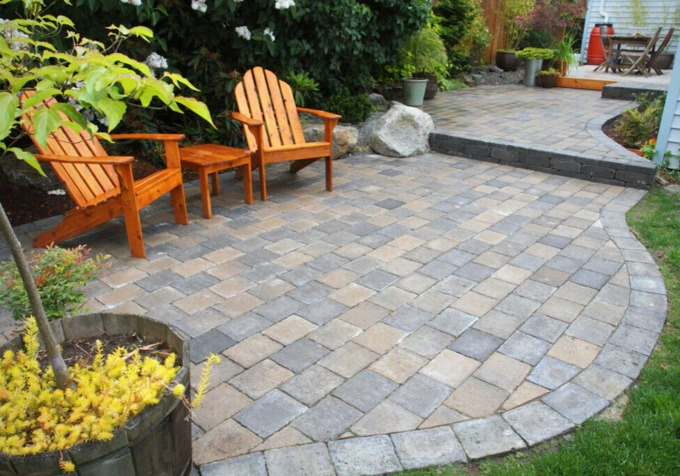 A patio with tan and gray stone pavers, two wooden Adirondack chairs, a small table, potted plants, and a raised dining area in the background.