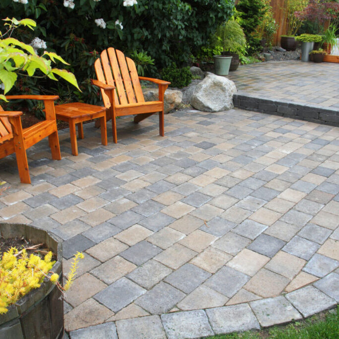 Two wooden chairs and a small table sit on a paved patio in a backyard garden, surrounded by plants and bordered by a raised stone platform.