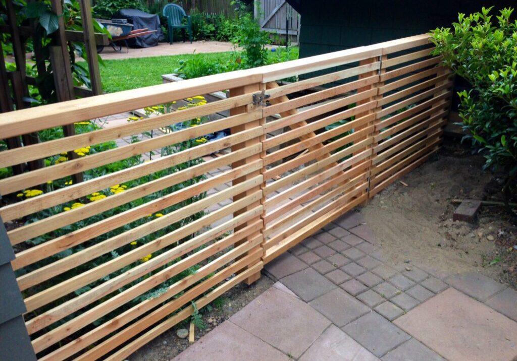 A horizontal wooden slat fence with a gate stands between a garden area with grass and flowers and a paved walkway.