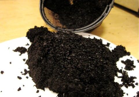 Coffee grounds spilled onto a white plate with a black coffee filter container tipped over in the background.