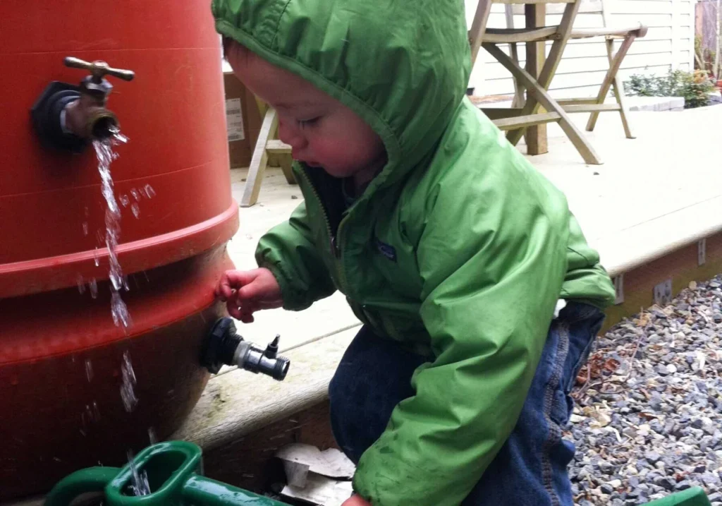 A young child in a green jacket is turning the spigot of a large red water barrel, watching water flow out onto the ground.