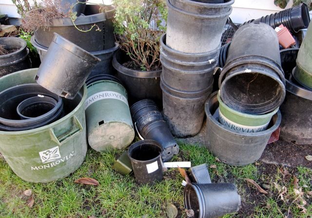 A pile of empty black and green plastic plant pots, some labeled "Monrovia," stacked and scattered on grass near a wall.