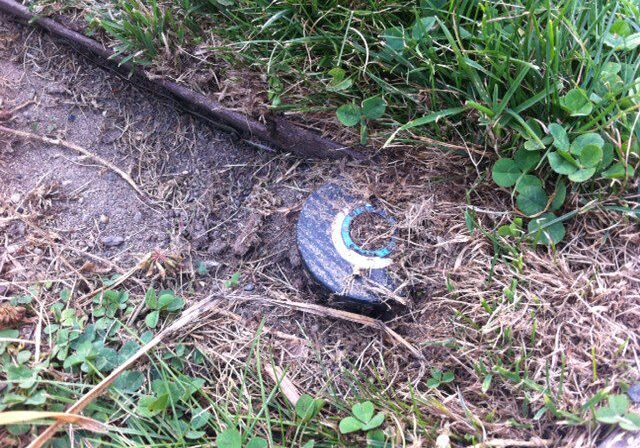 A partially buried, round irrigation sprinkler head is visible among grass and dirt, with the top marked by a worn white curved line.