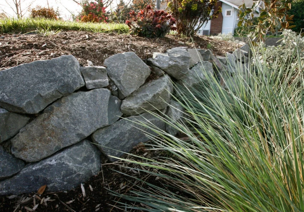 A stone retaining wall supports a sloped garden with ornamental grasses and mulch, with a house partially visible in the background.